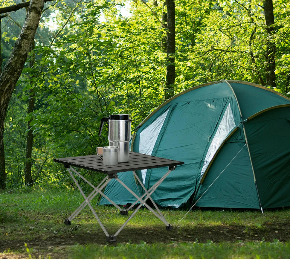 Camping scene with a teal tent and a folding table from The Adirondack Tool Co. with camping gear in a forest.