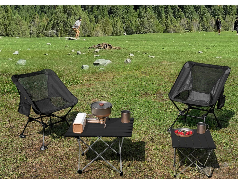 Two black folding camping tables from The Adirondack Tool Co. with chairs on a grassy field with trees in the background.