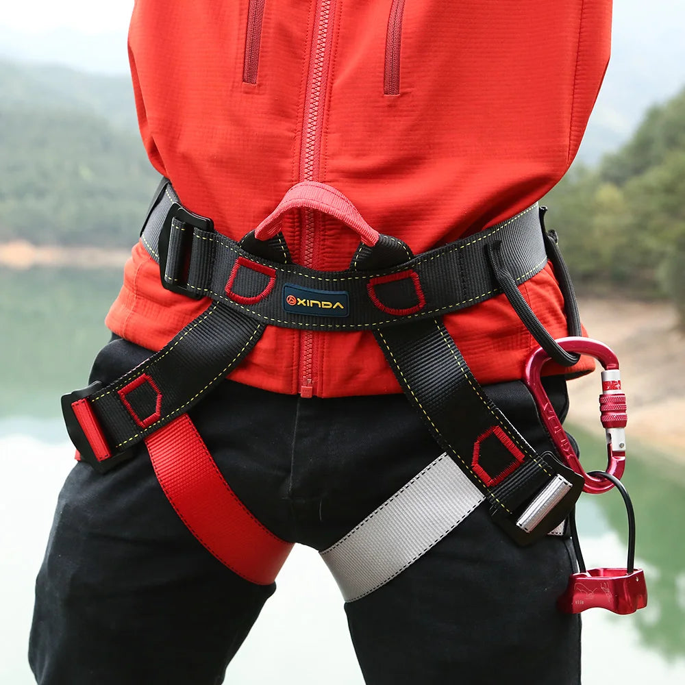 Person wearing a red jacket and black and red climbing harness with carabiners against a natural background - offered by the best climbing gear store in America, The Adirondack Tool Co.