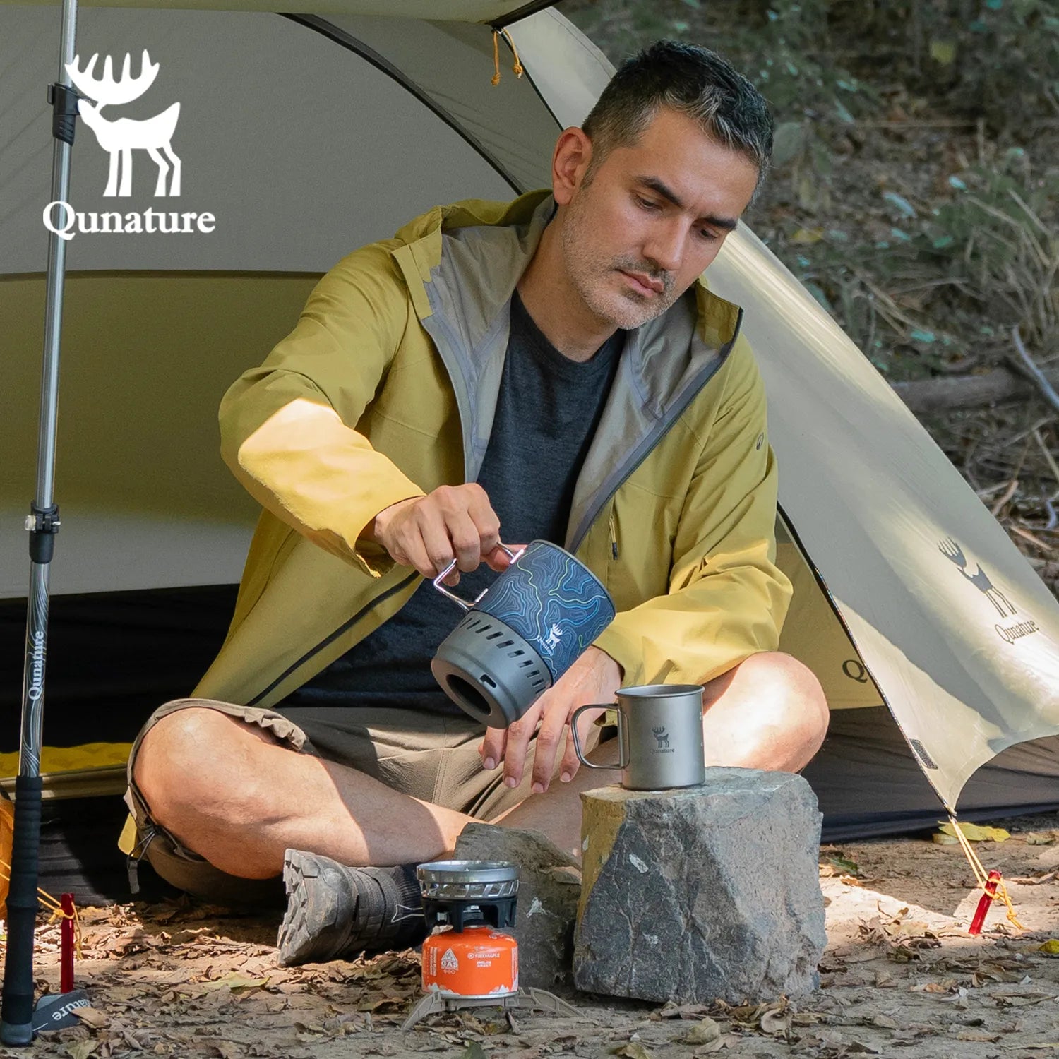 Man camping, preparing a drink using a portable stove and cookware in front of a tent with Qunature branding - offered by the best camping gear store in New York, The Adirondack Tool Co.